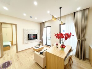 Uma sala de estar moderna com televisão na parede, mesa de jantar de madeira com cadeiras, um sofá pequeno e uma janela grande com cortinas.