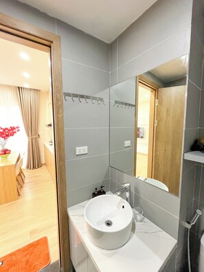 Um banheiro moderno com pia branca, espelho e azulejos cinzentos. Uma porta de madeira dá acesso a uma sala de jantar com mesa e cadeiras.