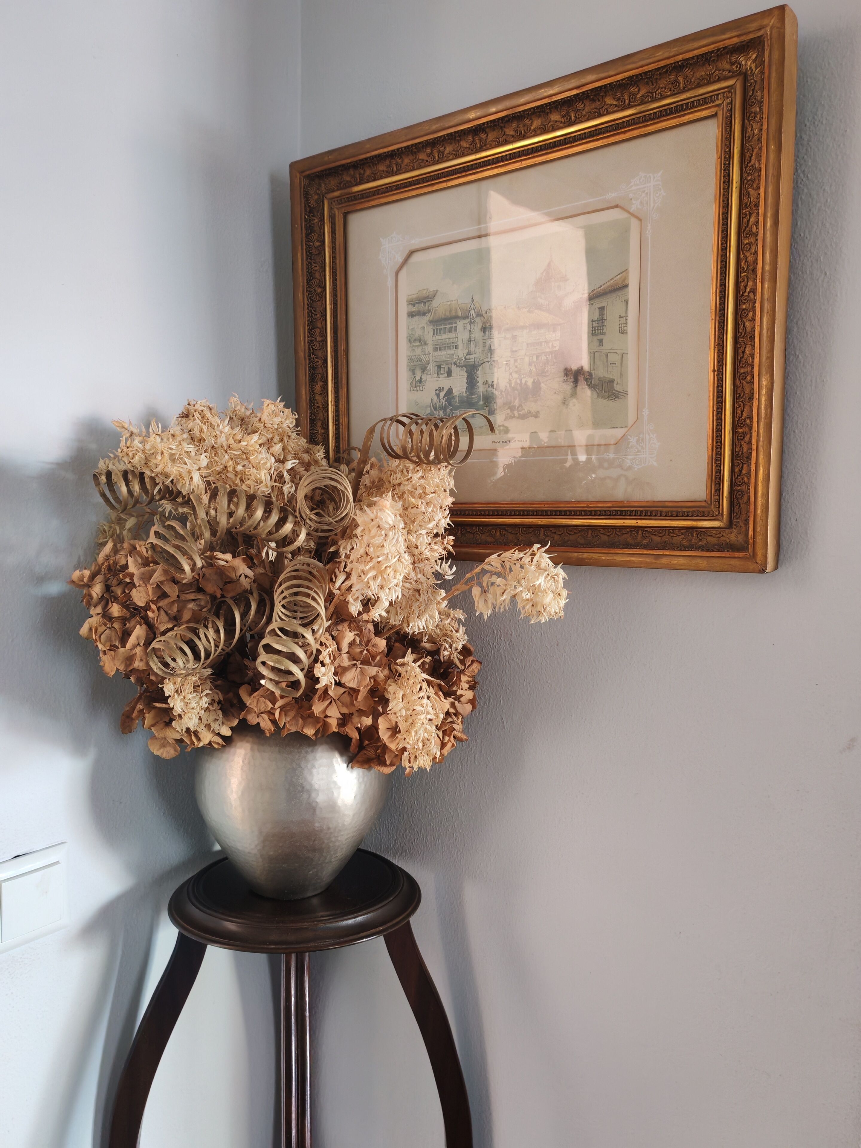 A silver vase with dried flowers on a dark wooden stool against a light wall, with a framed picture above.