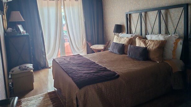 Une chambre à coucher avec un grand lit, une table de chevet avec une lampe, une chaise et une vue sur l’extérieur à travers une fenêtre avec des rideaux.