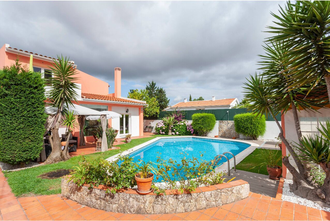 Una piscina rodeada por un jardín con diversas plantas y una casa con techo de tejas.