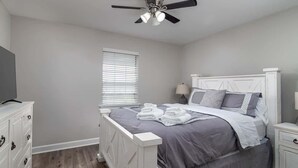 Une chambre à coucher comprenant un lit, un ventilateur de plafond, une commode, une table de chevet et une fenêtre avec des stores.