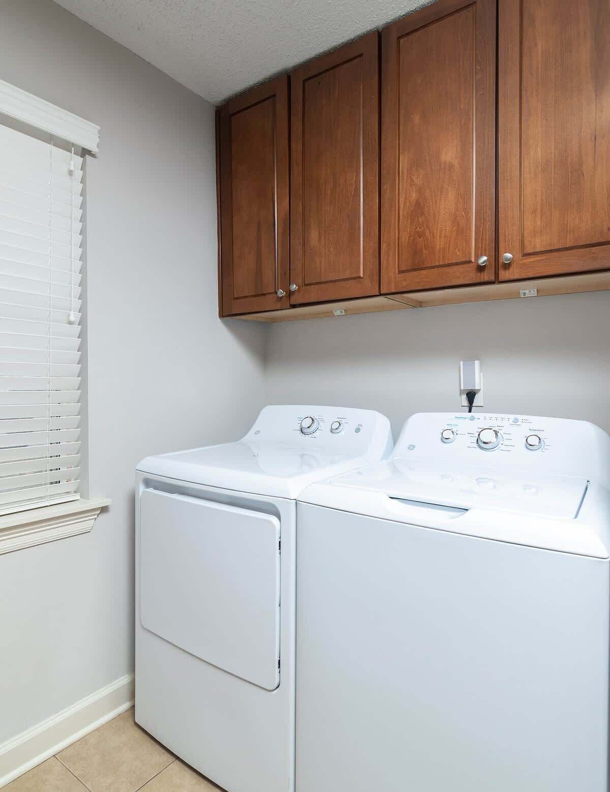 A laundry room with two white dryers, wooden cabinets above, and a window with blinds.