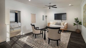 A bedroom with a bed, a desk, two chairs, a ceiling fan, a TV, and a potted plant.