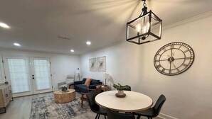 A modern living room with a round dining table, black chairs, a coffee table, a clock on the wall, and a hanging light fixture. - The Corner at Old Taylor Place (Oxford)