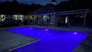 A swimming pool with blue lighting, a covered patio area, and a building with a chimney in the background. - The Corner at Old Taylor Place (Oxford)