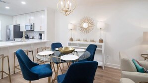 Una cocina moderna con armarios blancos, un refrigerador de acero inoxidable y un microondas. Hay un comedor con una mesa de vidrio y sillas azules.