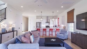 A modern living room with a sofa, coffee table, and striped armchair. A kitchen with red bar stools and a large TV is visible in the background. - The Lamar Landing (Oxford)