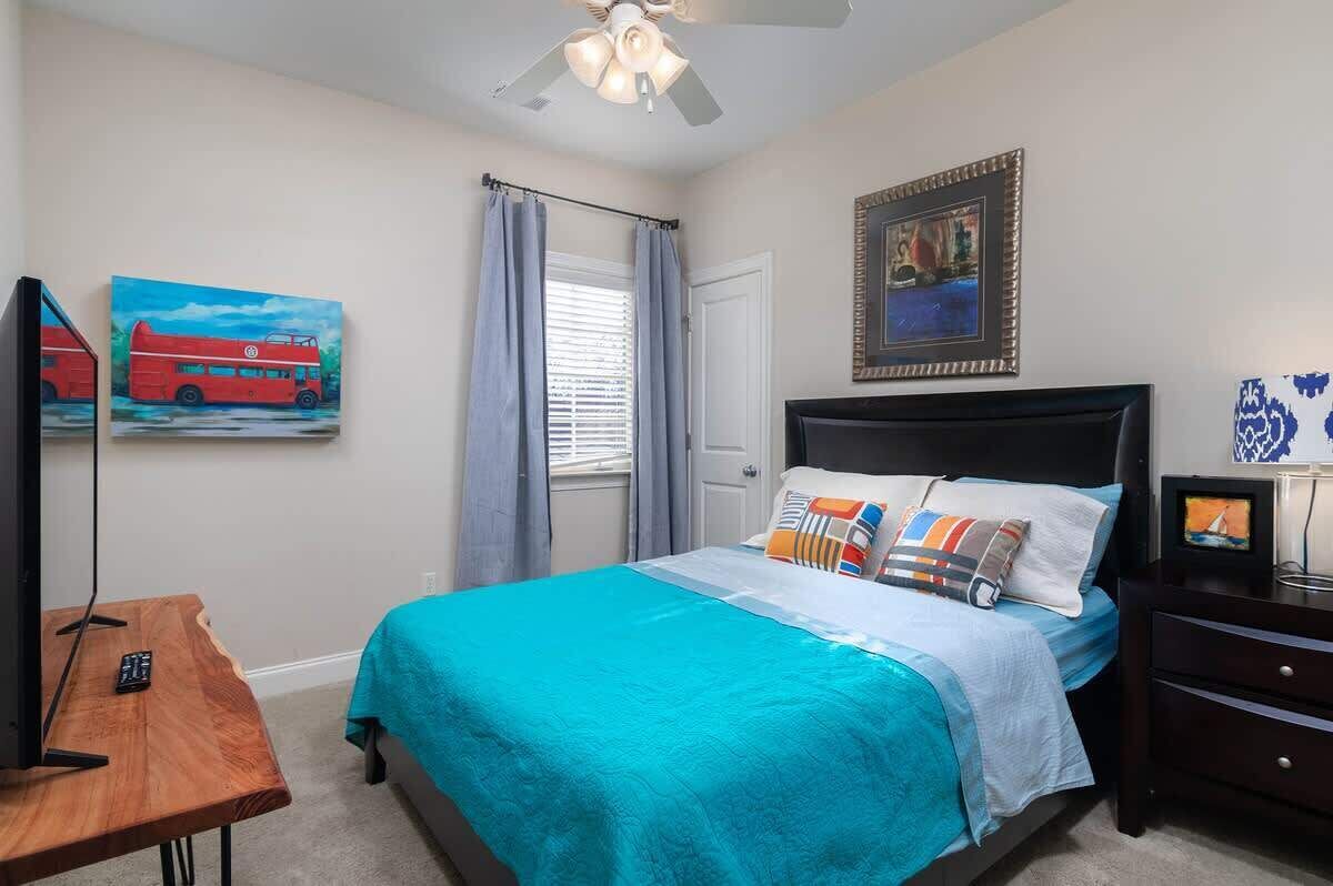 A bedroom with a bed, a desk, a ceiling fan, a window with curtains, and artwork on the walls.