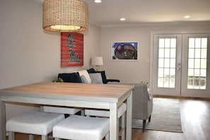 Een eethoek met een houten tafel en bankjes, een geweven lamp, een bank met kussens, een ingelijst beeld en een deur met glaspanelen.