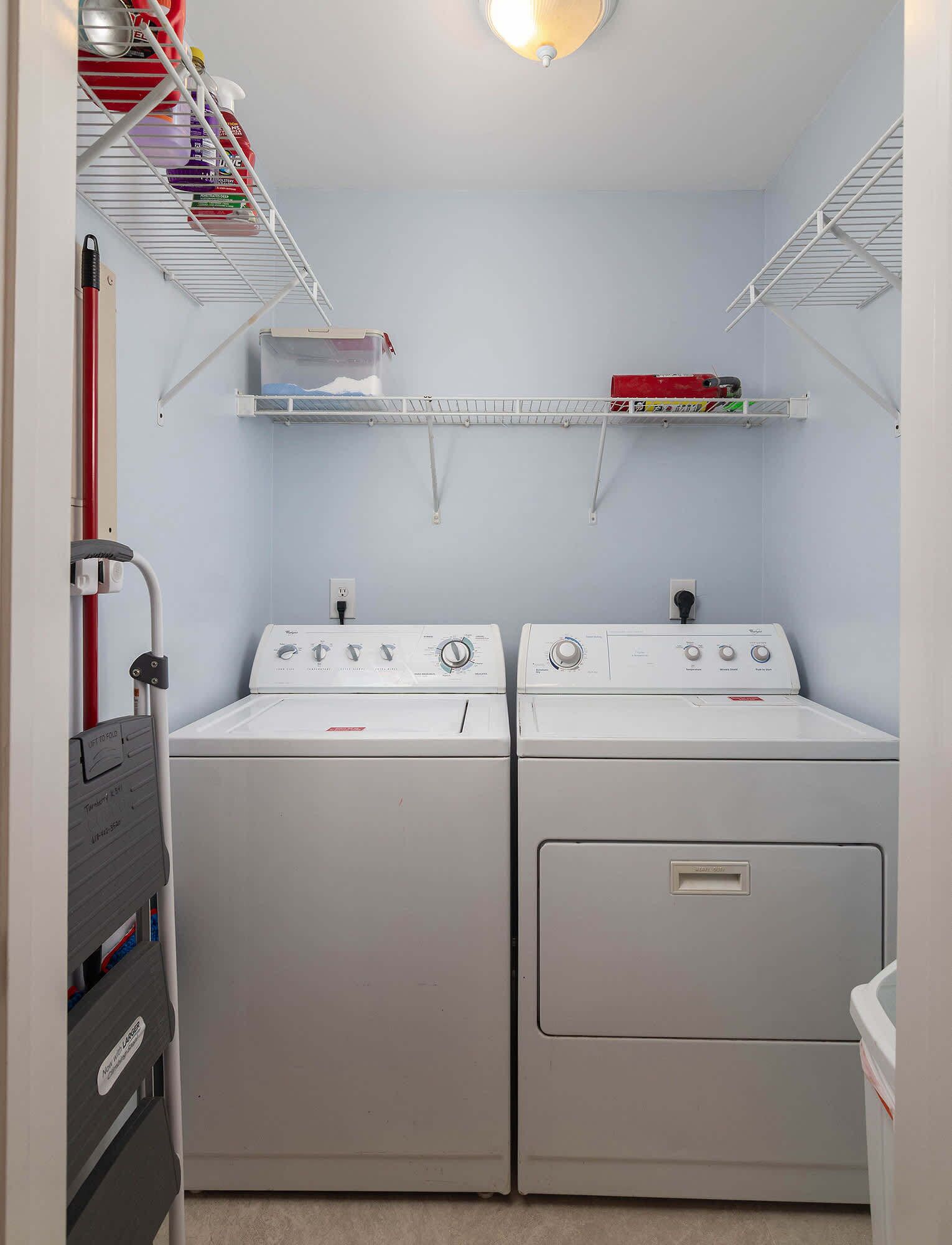 A laundry room with two front-loading washing machines, a drying rack, and shelves above for storage.