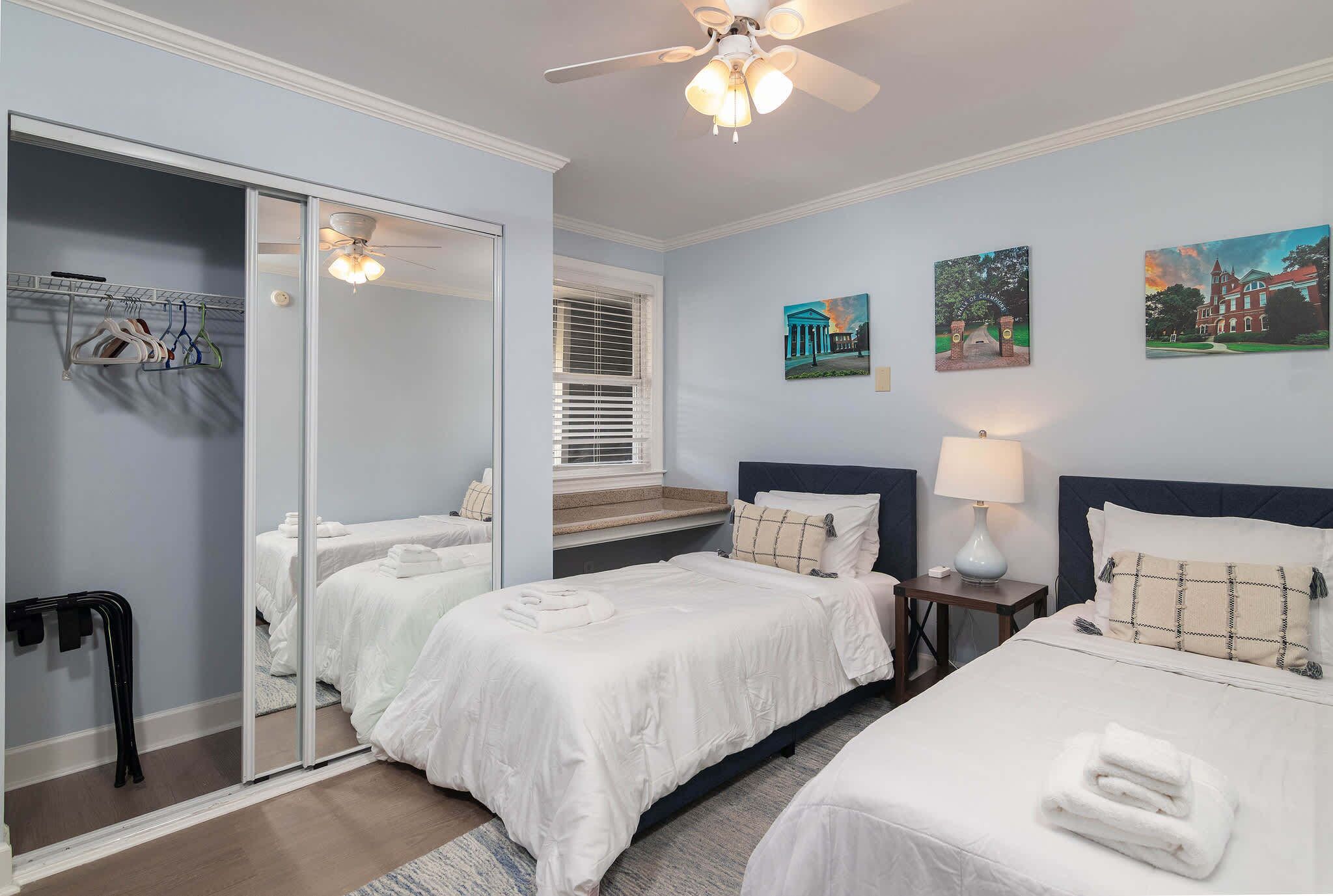 A hotel room with two beds, a mirror, a ceiling fan, and artwork on the walls.