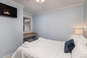 A bedroom with a bed, a TV, a window with blinds, a bedside lamp, and a small shelf. - Gated Condo, Close to Ole Miss! (Oxford)