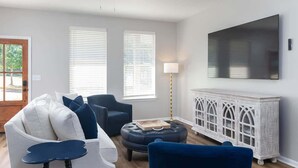 Ein Wohnzimmer mit einer Couch, Sesseln, einem Hocker, einem Fernseher und einem weißen Schrank.