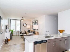 A modern kitchen with a dishwasher, a sink, and a countertop. There is a dining area with a table and chairs, and a living room with a sofa and a lamp. - The Middle Ground (Oxford)
