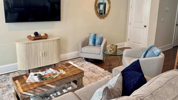 Una sala de estar con televisor de pantalla plana, una mesa de centro redonda, un sofá, un sillón, una mesita auxiliar pequeña y un aparador.