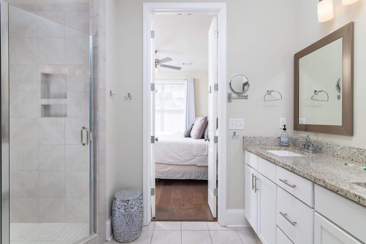 A bathroom with a shower, a sink, a mirror, and a bed visible through an open door.
