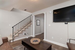 A living room with a staircase, a flat-screen TV mounted on the wall, and a coffee table with a basket. - Oxford Condo - Walk to Campus (Oxford)