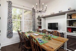 A dining room with a wooden table, chairs, a chandelier, and a television on a shelf. - Retreat in Blue (Oxford)