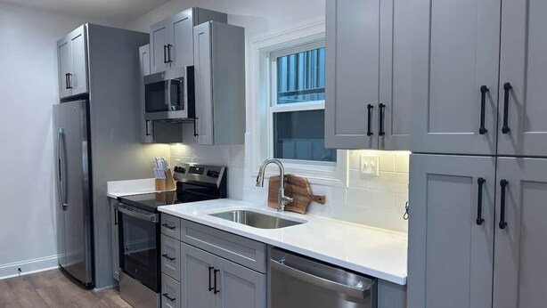 A modern kitchen with stainless steel appliances, white cabinetry, and a window with blue curtains. - The Rebel Sole (Oxford)
