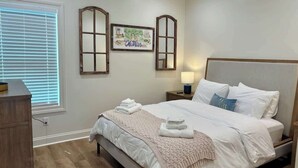 Ein Schlafzimmer mit Bett, Nachttisch, Lampe, Kommode und einem Fenster mit Jalousien.
