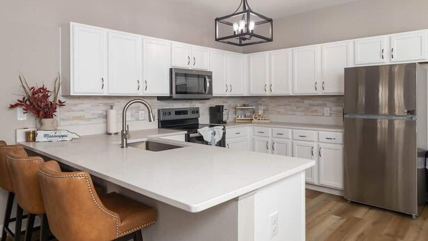 A modern kitchen with white cabinets, stainless steel appliances, a central island with bar stools, and a hanging light fixture. - The Modern Sip (Oxford)