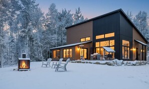 A modern two-story cabin with a fireplace, outdoor seating, and a view of a snowy forest. - Comfort & Spa Near the River | Chalet Le 3751 (Saint-Côme)