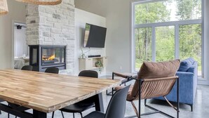 Uma sala de estar moderna com lareira de pedra, uma janela grande com vista para uma floresta e um espaço de jantar com mesa e cadeiras de madeira.