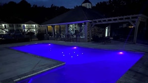 Ein Swimmingpool mit blauer Beleuchtung, ein überdachter Sitzbereich und im Hintergrund ein Gebäude mit einem Glockenturm.