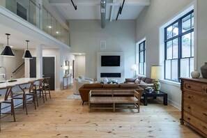 A spacious living area with a high ceiling, a large window, a flat-screen TV, a sofa, and a dining area with chairs.