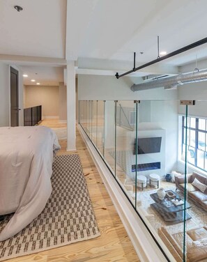 A modern bedroom with a large bed, a patterned rug, a glass railing, and a view of a living area with a sofa and a TV.
