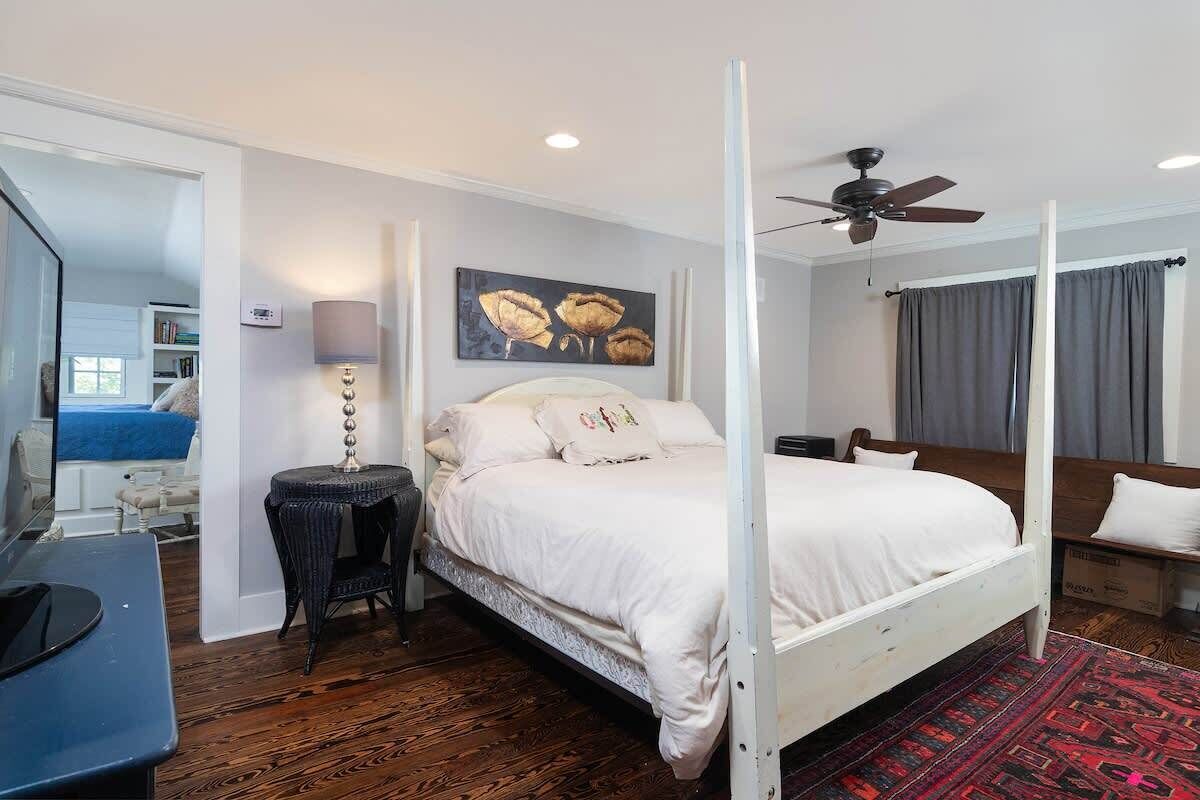 A four-poster bed with a white bedspread, a small side table with a lamp, a ceiling fan, and a painting on the wall.