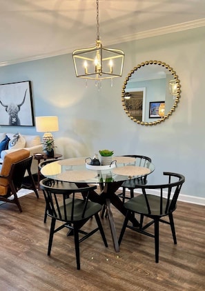 A dining area with a round glass table, black chairs, a chandelier, a mirror with a gold frame, and a wall-mounted lamp. - Oxford Contemporary Retreat (Oxford)