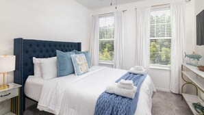 Une chambre à coucher avec une tête de lit capitonnée, un lit avec une literie blanche, des oreillers bleus et blancs, un plaid bleu et un ventilateur de plafond.
