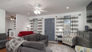 Una sala de estar con un sofá gris, una butaca roja, un sillón estampado, un mueble gris y un ventilador de techo.