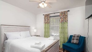 Un dormitorio con cama, una silla, ventilador de techo y cortinas.