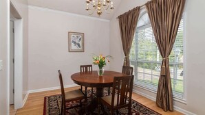 Comedor con una mesa redonda, sillas, una araña, una ventana con cortinas y un cuadro en la pared.