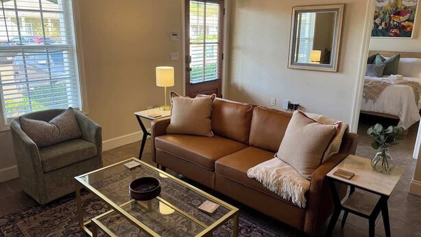 Ein Wohnzimmer mit braunem Ledersofa, einem Glastisch, einem gemusterten Teppich, einem Beistelltisch mit Lampe und einem Gemälde an der Wand.