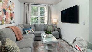 Een woonkamer met een grijze bank, een salontafel, een aan de muur bevestigde televisie en een raam met gordijnen.