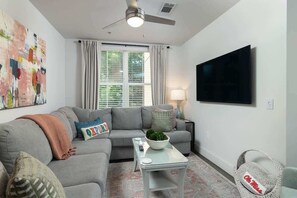 A living room with a grey sofa, a coffee table, a TV mounted on the wall, and a window with curtains.