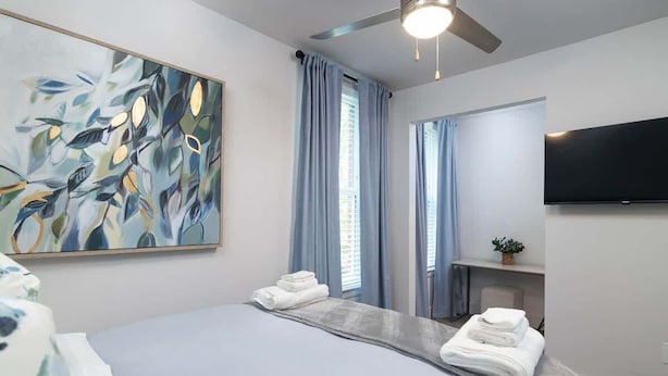 Een hotelkamer met een bed, een plafondventilator, een schilderij, een televisie en een raam met gordijnen.
