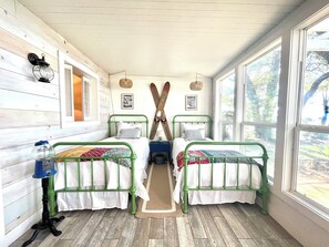 A cabin with two green metal beds, a wooden floor, a window with a view of trees, and a wall-mounted lantern. - Cozy Cottage on the Bay: Where memories are made, Private dock, and Sunset Views (Gulf Breeze)