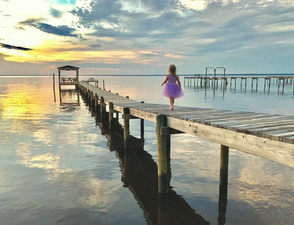 A wooden pier extending into calm water with a person in a purple dress walking away from the camera. - Cozy Cottage on the Bay: Where memories are made, Private dock, and Sunset Views (Gulf Breeze)