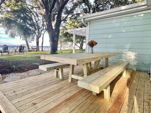 A wooden deck with a picnic table and benches, surrounded by trees and a view of the ocean. - Cozy Cottage on the Bay: Where memories are made, Private dock, and Sunset Views (Gulf Breeze)