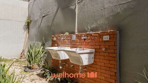 Ein kleiner Außenbereich mit zwei weißen Spülbecken, die an einer Ziegelwand angebracht sind, umgeben von Pflanzen und einer Betonwand.