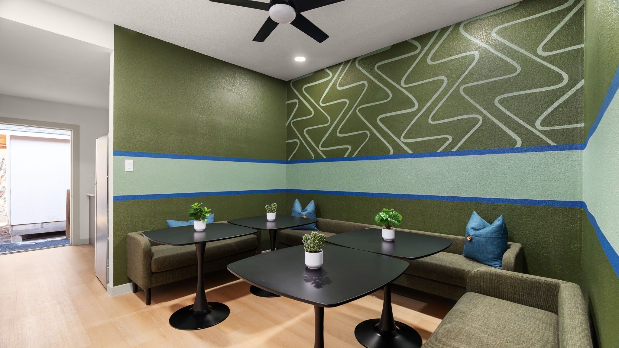 A room with a green and blue wall design, a ceiling fan, and a seating area with tables and chairs.