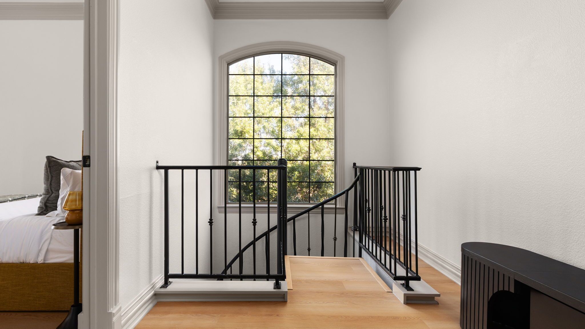 A staircase with black railings leading to a landing, a large arched window with a view of trees, and a bed with white bedding and a lamp.