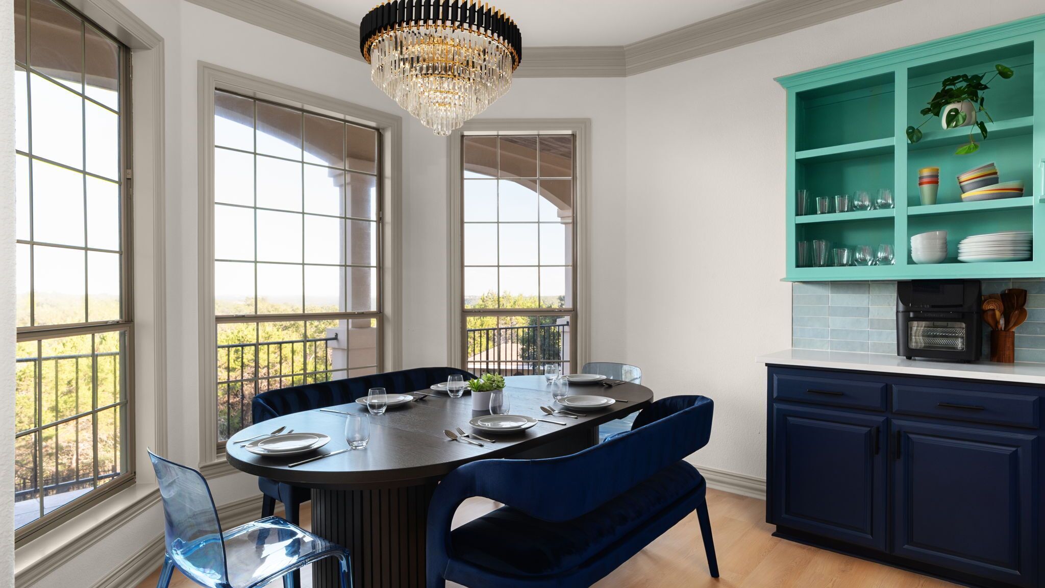 A dining area with a round table, blue chairs, and a chandelier. There are large windows with a view of greenery outside.