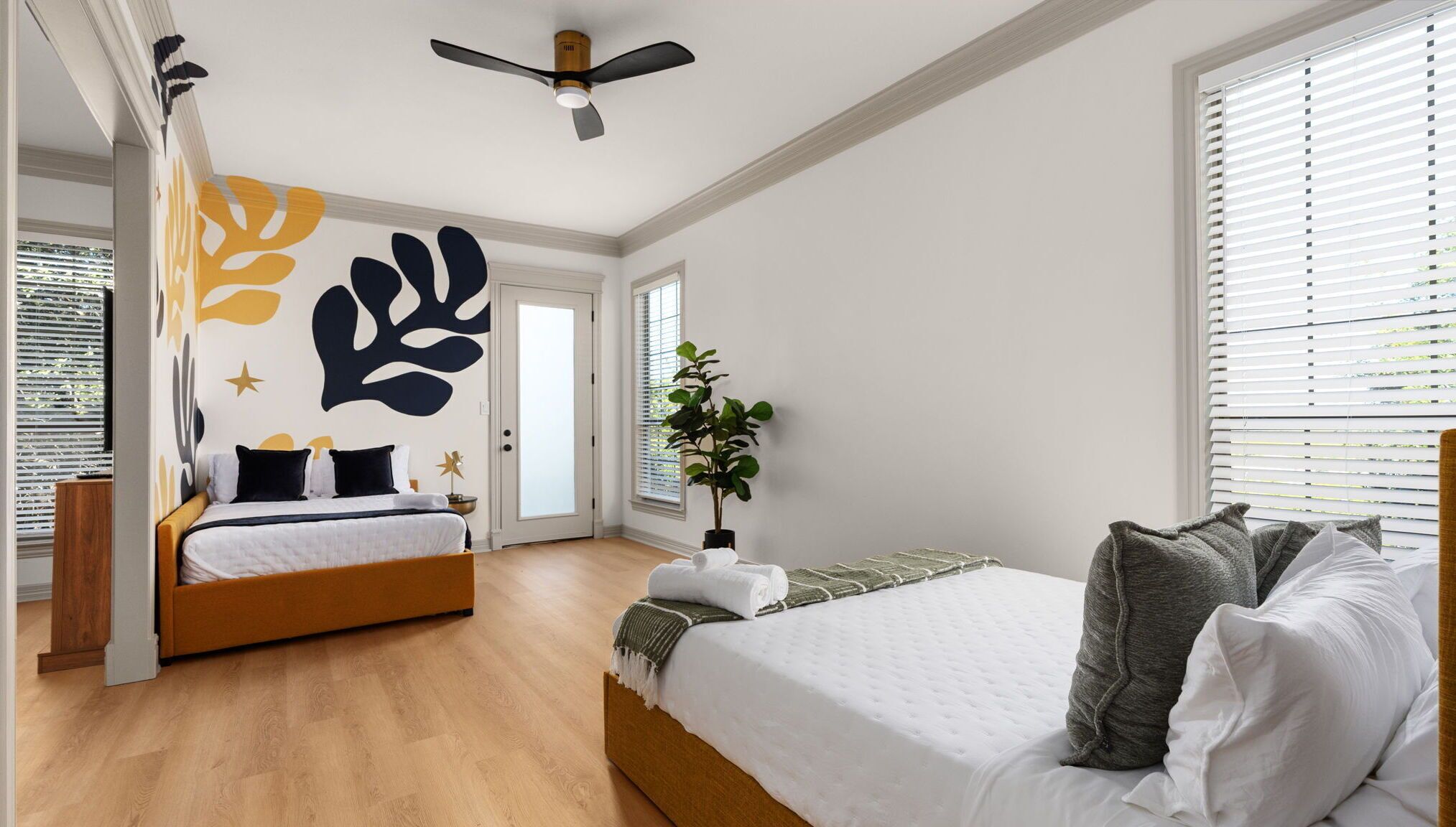 Ein Schlafzimmer mit einem Bett, einem zusätzlichen Bett, einem Deckenventilator, einer Topfpflanze und einer Wand mit abstrakter Kunst.