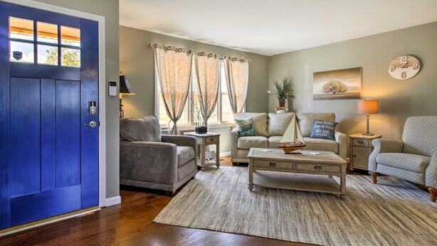 A living room with a blue door, a sofa, a coffee table, a patterned armchair, a clock, and a window with curtains.